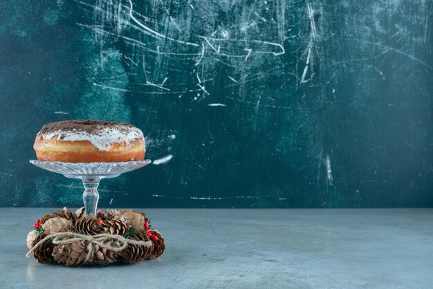 Glasierter Donut auf einem Glassockel inmitten eines Kiefernkranzes auf Marmor.