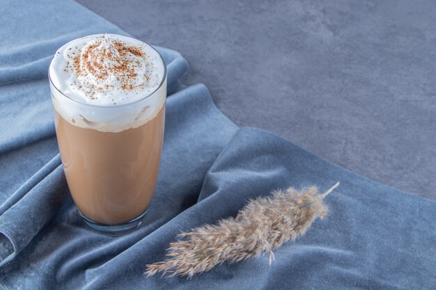 Glastasse Kaffee Latte auf einem Stück Stoff neben Pampasgras, auf blauem Hintergrund.