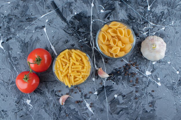 Glastassen ungekochte Nudeln mit zwei frischen Tomaten und Knoblauch auf einer Marmoroberfläche.