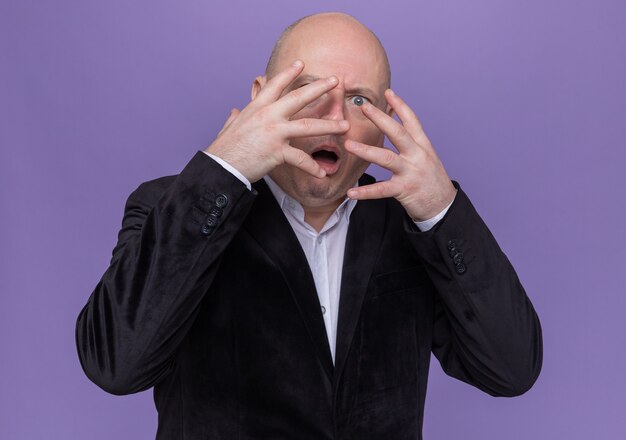 Glatzköpfiger Mann mittleren Alters im Anzug, der sein Gesicht mit Handflächen bedeckt, die durch Finger, die über lila Wand stehen, nach vorne schauen