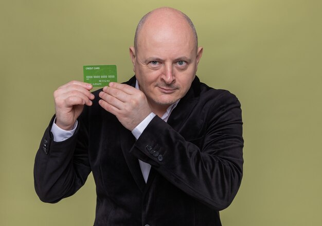 Glatzkopf mittleren Alters im Anzug, der schlau lächelnde Kreditkarte zeigt, die über grüner Wand steht
