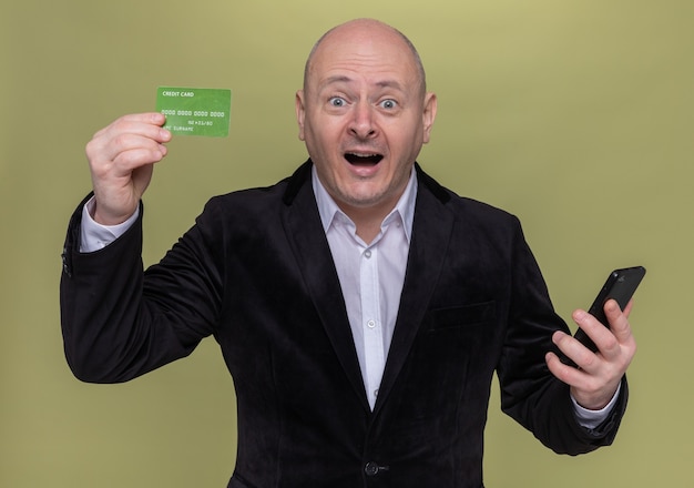 Glatzkopf mittleren Alters im Anzug, der zeigt, dass Kreditkarte überrascht und glücklich über grüner Wand steht