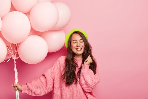 Glücklich erfreut brünette asiatische Dame steht mit Luftballons, genießt coole Party mit Freunden, trägt Baskenmütze und losen Pullover, feiert Jubiläum