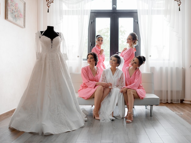 Glücklich gelächelte Brautjungfern mit Braut schaut auf das Hochzeitskleid im hellen Raum, Hochzeitsvorbereitung