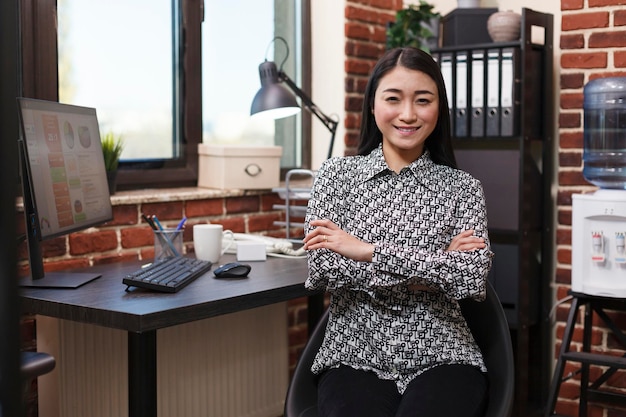 Glücklich lächelnde asiatische Geschäftsfrau, die am Schreibtisch im Büro des Finanzunternehmens sitzt, während sie in die Kamera schaut. Fröhlicher Projektmanager, der im Inneren einer modernen Marketingagentur sitzt.