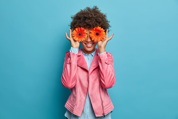 glücklich lächelnde Frau versteckt Gesicht mit zwei orange Gerbera, mag Blumen, drückt Glück und Freude aus. Fröhlicher Florist, der hübschen Blumenstrauß zum Verkauf machen wird, arbeitet im Blumenladen