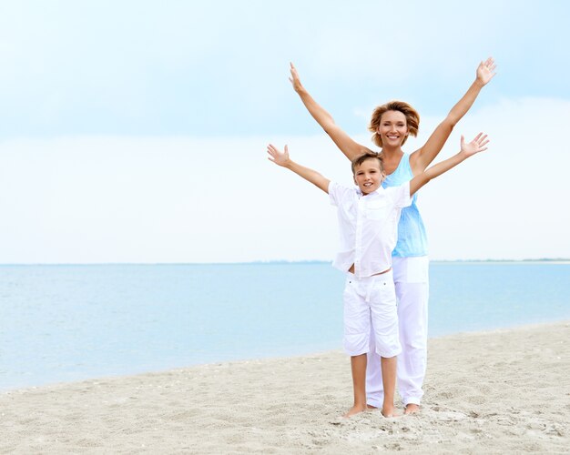 Glücklich lächelnde Mutter und Sohn, die am Strand mit erhobenen Händen stehen.