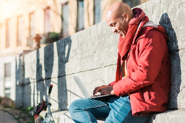 Glücklich lächelnder hispanischer Mann mit einem Laptop-Computer, während er sich an einer Wand in einer Straße von Lyon lehnt