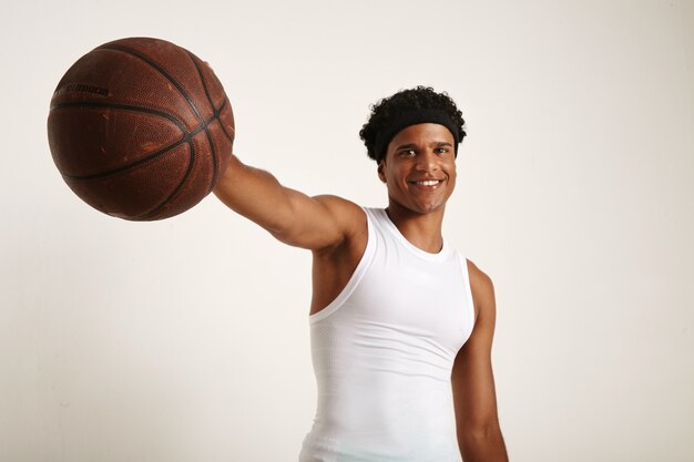 glücklich lächelnder junger schwarzer Athlet mit Afro und Stirnband im weißen Hemd, das einen Vintage braunen Basketball heraushält