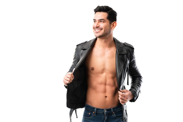 Glücklich lächelnder Mann in Lederjacke, der seine muskulösen Bauchmuskeln auf weißem Hintergrund zeigt