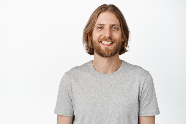 Glücklich lächelnder Mann mit blonden Haaren und Bart, Blick in die Kamera, stehend in grauem T-Shirt, isoliert auf weißem Hintergrund