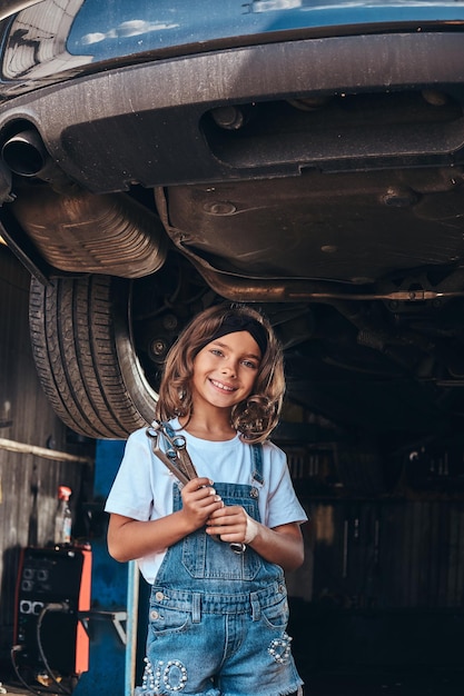 Glücklich lächelndes Mädchen steht unter dem Auto in der Autowerkstatt mit Schraubenschlüssel in den Händen.