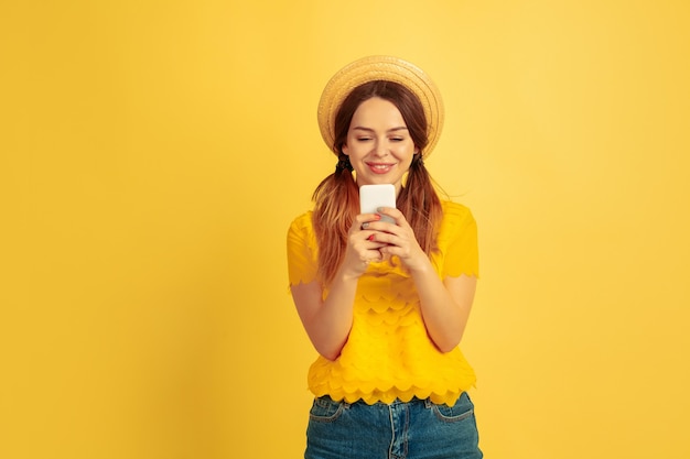 Glücklich mit dem Smartphone. Porträt der kaukasischen Frau auf gelbem Studiohintergrund. Schönes weibliches Modell im Hut. Konzept der menschlichen Emotionen, Gesichtsausdruck, Verkauf, Anzeige. Sommerzeit, Reisen, Resort.