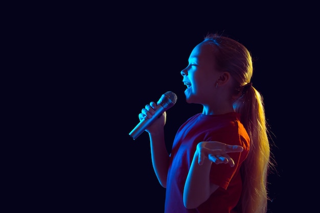 Glücklich singen. Porträt des kaukasischen Mädchens auf dunklem Studiohintergrund im Neonlicht. Schönes weibliches Modell mit Lautsprecher. Konzept der menschlichen Emotionen, Gesichtsausdruck, Verkauf, Anzeige, Hobby, Traum, Musik.