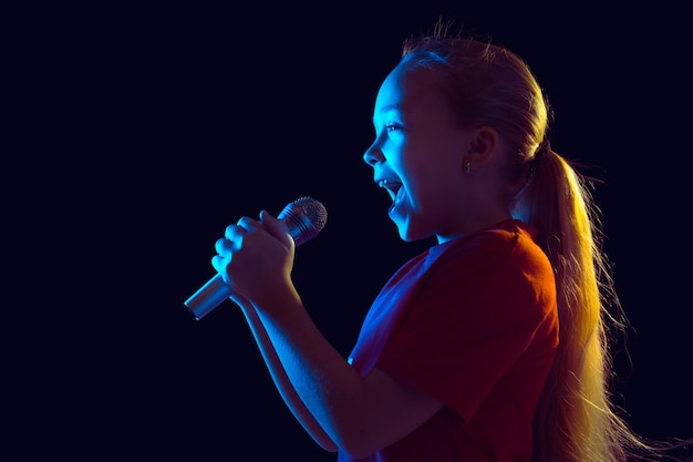Glücklich singen. Porträt des kaukasischen Mädchens auf dunklem Studiohintergrund im Neonlicht. Schönes weibliches Modell mit Lautsprecher.