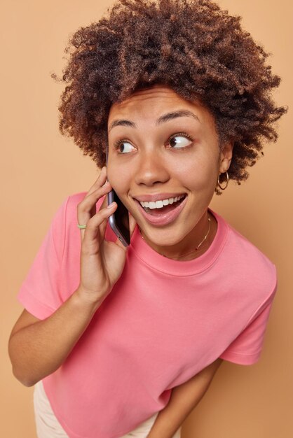 Glücklich überraschte junge Frau hat Telefongespräche, die sich mit frohem Ausdruck konzentrieren, verbringt Freizeit damit, mit Freunden über Handy zu klatschen, trägt rosa T-Shirt einzeln auf beigem Hintergrund