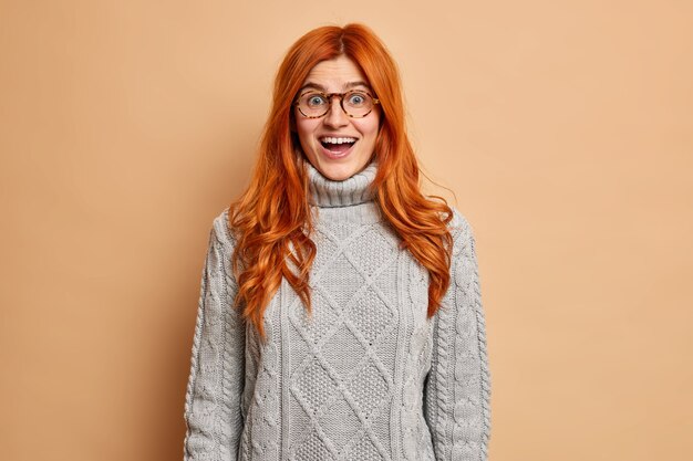 Glücklich überraschte rothaarige Frau sieht mit geöffnetem Mund aus und kann nicht an ihren plötzlichen Erfolg glauben, gekleidet in einen gestrickten Pullover.
