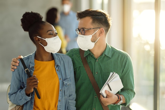 Glücklich umarmtes College-Paar mit schützenden Gesichtsmasken, das durch einen Flur geht