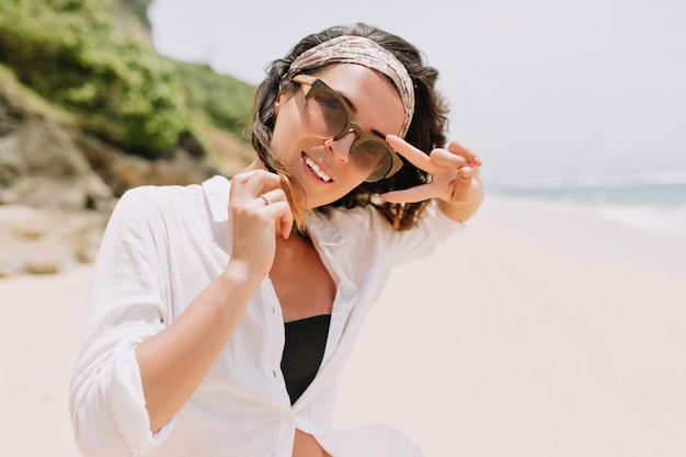 Glücklich verlassene junge Frau mit dunklem gewelltem Haar trägt schwarze Brille und Haarschmuck im weißen Hemd geht den weißen Sandstrand im Sonnenlicht hinunter