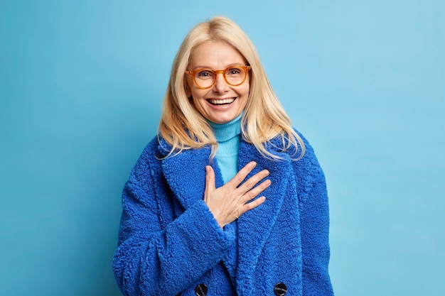 glückliche ältere blonde europäische Frau amüsiert von humorvollem Witz lacht positiv hält Hand auf Brust in winterblauem Mantel gekleidet.