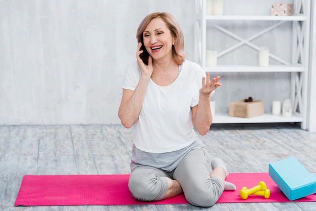 Glückliche ältere Frau, die auf dem Mobiltelefon sitzt auf Yogamatte spricht
