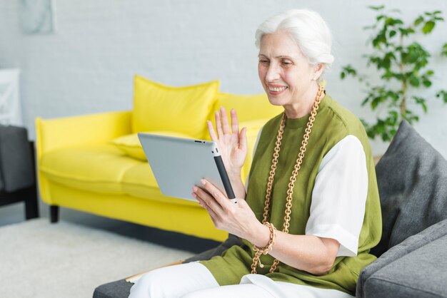 Glückliche ältere Frau, die auf dem Sofa betrachtet die digitale Tablette wellenartig bewegt ihre Hand sitzt