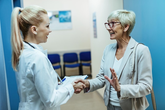 Glückliche ältere Frau, die einem Arzt die Hände schüttelt, während sie mit ihr auf dem Krankenhausflur spricht