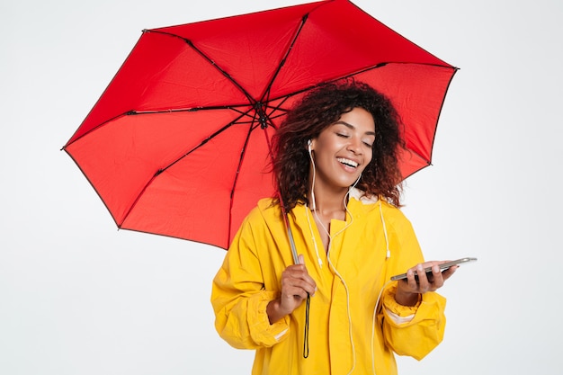 Glückliche afrikanische Frau im Regenmantel, der sich unter Regenschirm versteckt und Musik auf ihrem Smartphone über weißem Hintergrund hört