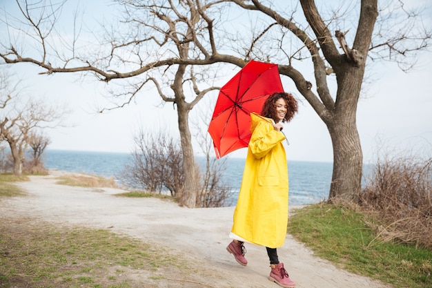 Glückliche afrikanische lockige junge Dame, die gelben Mantel hält Regenschirm trägt