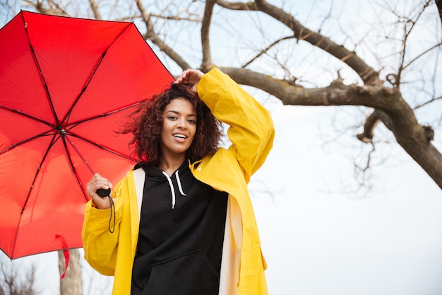 Glückliche afrikanische lockige junge Dame, die gelben Mantel mit Regenschirm trägt.
