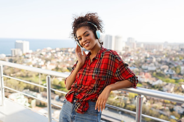 Glückliche afroamerikanische Frau, die reizende Musik durch Kopfhörer genießt, gekleidet im karierten Hemd, stehend auf Dach. Hintergrund der Stadtlandschaft.