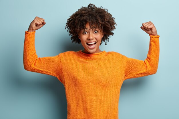 Glückliche afroamerikanische Frau mit lockigem Haar, hebt Hände und zeigt Muskeln, zeigt ihre Stärke, trägt orangefarbenen Pullover