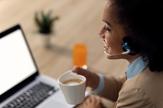 Glückliche afroamerikanische Geschäftsfrau trinkt Kaffee während der Telefonkonferenz über Laptop