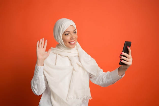 Glückliche arabische Frau im Hijab mit Handy, das selfie macht. Porträt des lächelnden Mädchens, das am roten Studiohintergrund aufwirft. Junge emotionale Frau. menschliche Emotionen, Gesichtsausdruck Konzept.