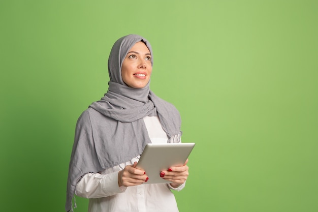 Kostenloses Foto glückliche arabische frau im hijab mit laptop. porträt des lächelnden mädchens, das am grünen studiohintergrund aufwirft. junge emotionale frau. menschliche emotionen, gesichtsausdruck konzept. vorderansicht.