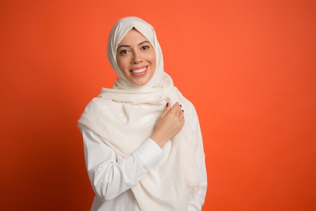 Kostenloses Foto glückliche arabische frau im hijab. porträt des lächelnden mädchens, das am roten studiohintergrund aufwirft. junge emotionale frau. die menschlichen emotionen, gesichtsausdruck konzept. vorderansicht.