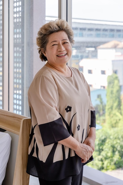 Glückliche asiatische ältere ältere Frau, die Stock hält, steht am Schlafzimmerfenster mit Blick auf die Stadt und blickt in die Kamera