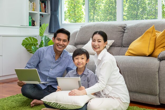 Glückliche asiatische Eltern sitzen zusammen mit Jungen auf dem Boden im Wohnzimmer Vater arbeitet am Laptop und Sohn mit Tablet, während die Mutter in die Kamera schaut