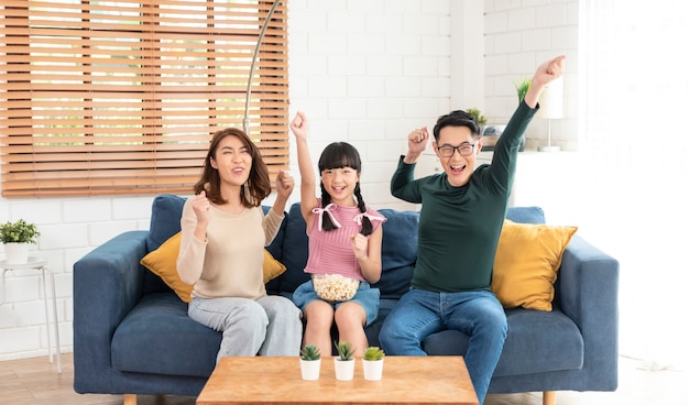 Glückliche asiatische Familie, die Popcorn isst und zusammen auf dem Sofa im Wohnzimmer fernsehen