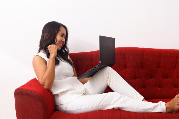 Glückliche asiatische frau, die einen laptop beim sitzen auf rotem sofa verwendet.