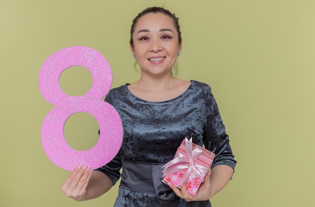 Kostenloses Foto glückliche asiatische frau, die nummer acht aus pappe und geschenk hält, die vorne lächelnd fröhlich den internationalen frauentag feiert, der über grüner wand steht