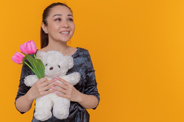 Glückliche asiatische Frau, die Strauß der rosa Tulpen und des Teddybären hält, der fröhlich beiseite schaut, der internationalen Frauentag feiert über orange Wand steht