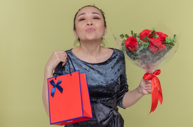 Glückliche asiatische Frau, die Strauß der roten Rosen und der Papiertüte mit Geschenk hält, das einen Kuss bläst, der internationalen Frauentag feiert, der über grüner Wand steht