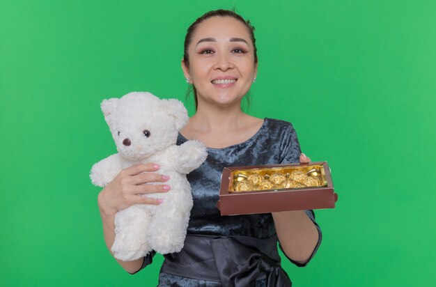 Glückliche asiatische Frau, die Teddybär und Schachtel Pralinen als ein Geschenk hält, das fröhlich den internationalen Frauentag feiert, der über grüner Wand steht