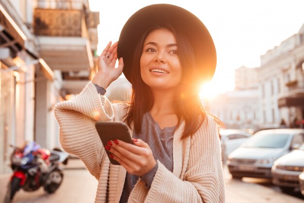 glückliche asiatische Frau in der Wolljacke, die ihren Hut berührt, während Handy in der Stadt im Freien hält