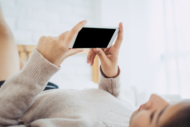 Glückliche asiatische Frauen benutzen intelligentes Telefon mit schwarzem leerem leerem Bildschirm auf dem Bett am Morgen