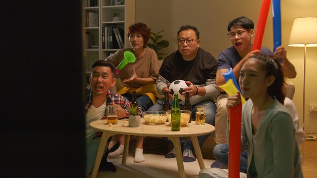 Glückliche asiatische freunde oder fußballfans, die fußball im fernsehen sehen und zu hause den sieg feiern