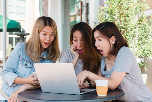 Glückliche asiatische Freundinnen der Gruppe, die Medieninhalt auf Linie im Laptop aufpassen, der in der Kaffeestube sitzt