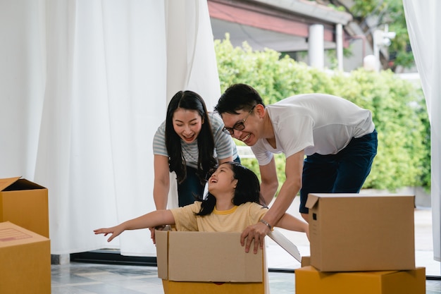 Glückliche asiatische junge familie, die den spaß lacht hat, in neues haus umzuziehen. japanische eltern bemuttern und der vater, der aufgeregtem reiten des kleinen mädchens helfend lächelt, das in der pappschachtel sitzt. neues eigentum und umzug.