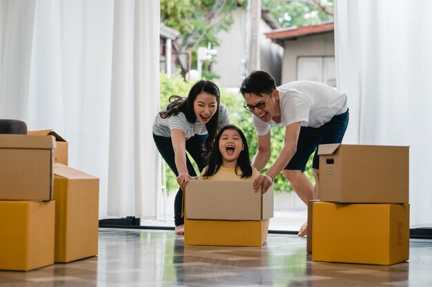 Glückliche asiatische junge Familie, die den Spaß lacht hat, in neues Haus umzuziehen. Japanische Eltern bemuttern und der Vater, der aufgeregtem Reiten des kleinen Mädchens helfend lächelt, das in der Pappschachtel sitzt. Neues Eigentum und Umzug.